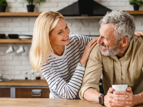 An older couple smiling at each other while in a kitchen.