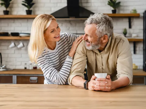 An older couple smiling at each other while in a kitchen.