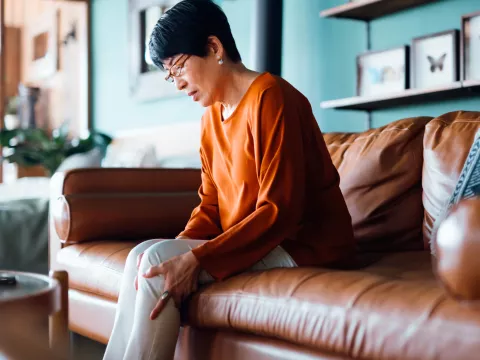 A woman sits on a couch and hold her hand to her leg.