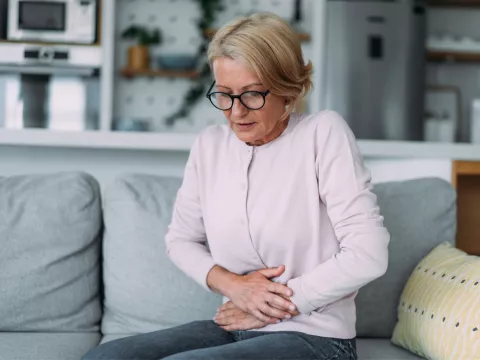 A senior woman sitting on a couch holding her stomach in discomfort while at home.