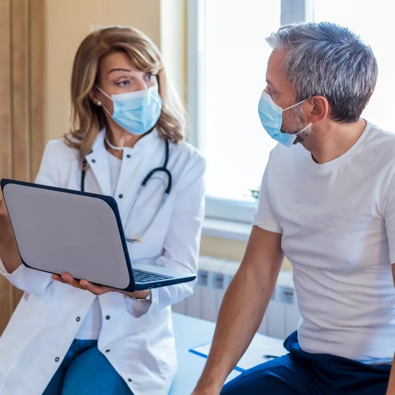 Female doctor talking with male patient holding laptop, both masked