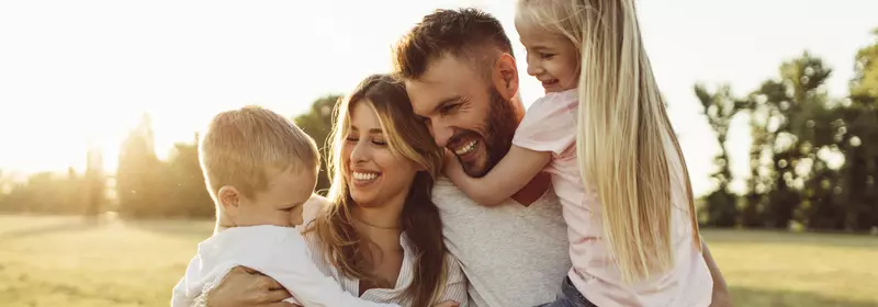 A family of four, the mother holding the son and the father holding the daughter, all laughing and smiling. The sun setting behind them.