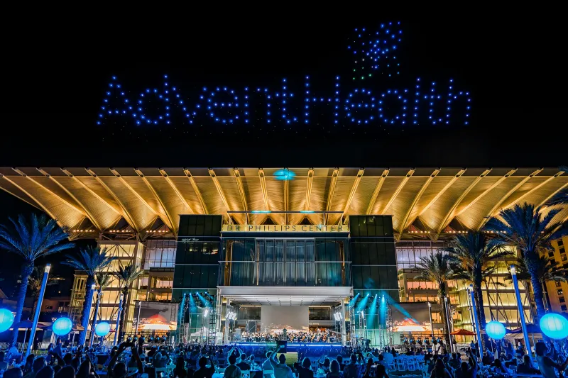 AdventHealth Orchestra performing at a night outdoor concert at Dr. Phillips Center for the Performing Arts with drones making the AdventHealth logo in the sky.